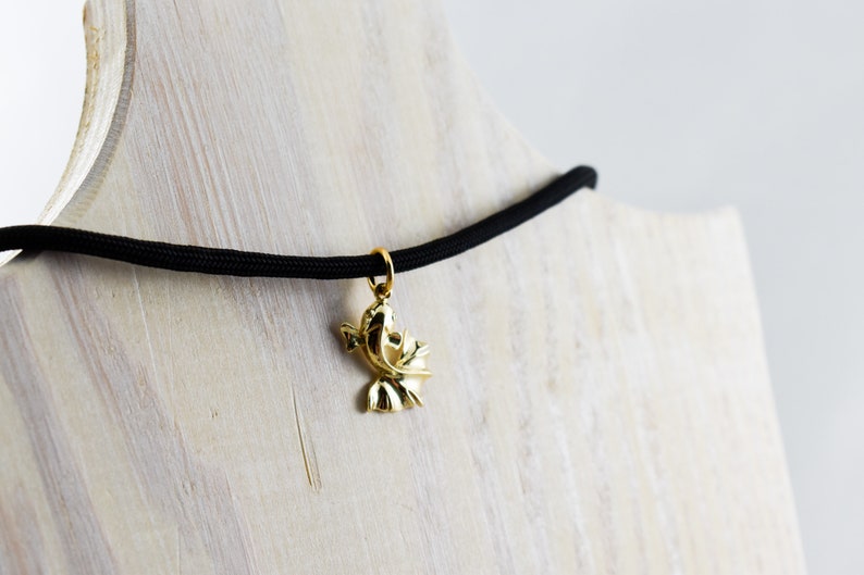 Puede incluir: Un collar con un colgante de p&aacute;jaro dorado en un cord&oacute;n el&aacute;stico negro. El collar se muestra sobre una superficie de madera.