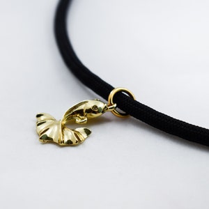 Puede incluir: Un colgante de pez dorado cuelga de un collar de cord&oacute;n negro. El colgante est&aacute; detallado con una aleta y un ojo.