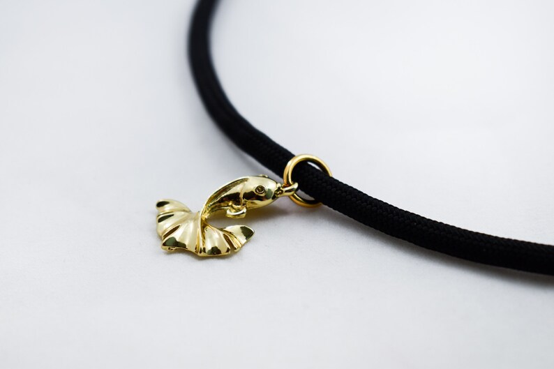 Puede incluir: Un colgante de pez dorado cuelga de un collar de cord&oacute;n negro. El colgante est&aacute; detallado con una aleta y un ojo.
