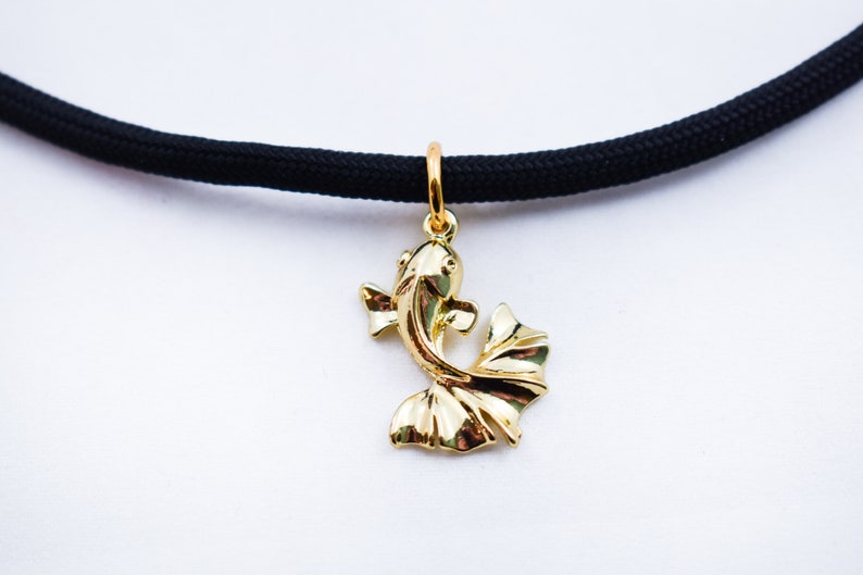 Puede incluir: Un colgante de pez dorado cuelga de un collar de cord&oacute;n negro. El pez est&aacute; estilizado con un cuerpo curvo y una gran aleta caudal que fluye.