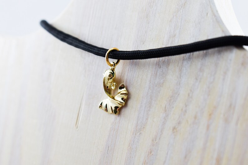 Puede incluir: Un collar con un colgante de pez dorado en un cord&oacute;n negro. El colgante es un pez estilizado con una cola y aletas curvas.