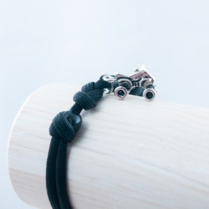 May include: Black paracord bracelet with a silver monster truck charm. The bracelet is tied with a knot and has a black bead.