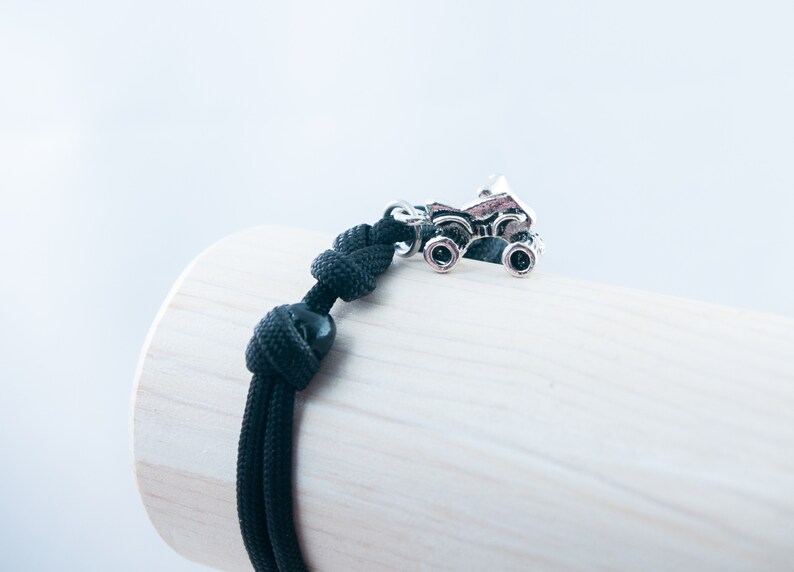 May include: Black paracord bracelet with a silver monster truck charm. The bracelet is tied with a knot and has a black bead.