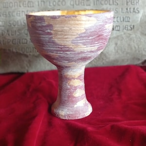 May include: A handcrafted chalice with a wide bowl and a slender stem. The chalice is painted with a maroon and gold color scheme, with visible brushstrokes. The interior of the chalice is gold. The chalice is set against a red background.