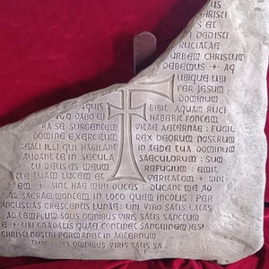 Könnte beinhalten: Eine graue Steintafel mit eingraviertem lateinischen Text und einem Kreuzdesign. Der Stein hat eine unregelmäßige Form und ist auf einem Holzständer montiert. Der Text ist in einer klassischen Schriftart.