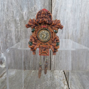 May include: A small wooden cuckoo clock with a carved face and green leaves. The clock face has Roman numerals and a gold-colored hand. The clock is hanging from a chain with two pine cone-shaped weights.