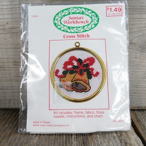 May include: A cross stitch kit featuring a framed design of two gold bells with a red bow. The kit includes a frame, fabric, floss, needle, instructions, and a chart. The packaging is white with green and red accents. The text "Santa's Workbench" and "Cross Stitch" are printed on the packaging. The kit is made in Taiwan and is copyright 1988, Fabri-Centers of America, Inc.