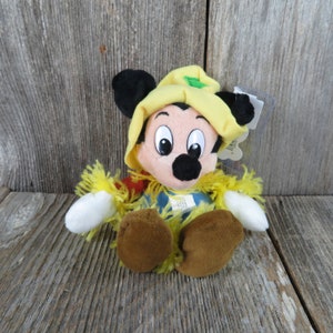May include: A plush Mickey Mouse doll wearing a yellow hat and a yellow straw-like collar. The doll has black ears, a black nose, and brown feet. The doll is sitting on a wooden surface.