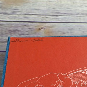 May include: A red hardcover book with a blue spine. The book has a white line drawing on the cover and the name "Sharon Todd" is written in black ink on the front cover.