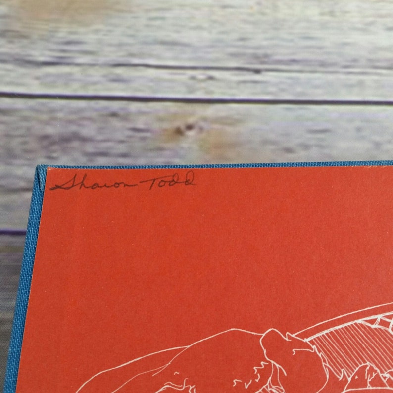 May include: A red hardcover book with a blue spine. The book has a white line drawing on the cover and the name "Sharon Todd" is written in black ink on the front cover.