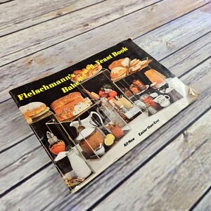 May include: A vintage cookbook titled "Fleischmann's Bake Yeast Book" with a black cover and yellow text. The cover features images of baked goods and ingredients. The book is on a light wood surface.