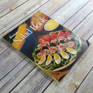 May include: A vintage Sunset Salad Book cookbook featuring a colourful image of a salad on the cover. The salad includes shrimp, olives, tomatoes, green beans, and hard-boiled eggs.