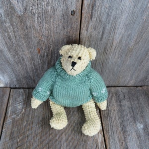 May include: A knitted cream-colored teddy bear wearing a green sweater with white snowflake accents. The bear is sitting on a wooden surface.