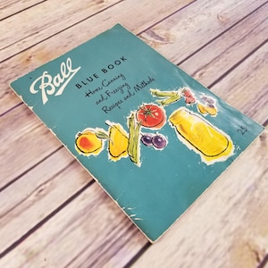 Könnte beinhalten: Ein blaues Vintage-Kochbuch von Ball Blue Book mit dem Titel "Home Canning and Freezing Recipes and Methods" und einem Preis von 25 Cent. Der Umschlag zeigt eine farbenfrohe Illustration verschiedener Früchte und Gemüse, darunter eine Tomate, Birnen, Pfirsiche, grüne Bohnen und Blaubeeren, die alle um ein klares Glasgefäß angeordnet sind.