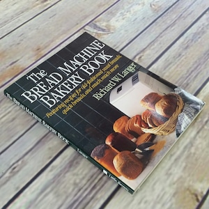 May include: A green and black book titled "The Bread Machine Bakery Book" by Richard W. Langer. The book features recipes for old-fashioned yeast breads, quick breads, and more. The cover features a bread machine and a basket of freshly baked bread.