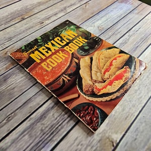 May include: A vintage cookbook titled "Sunset Mexican Cook Book" with a cover featuring images of Mexican food. The cover displays a bowl of salsa, a plate of tacos, and a bowl of beans. The book is on a wooden surface.