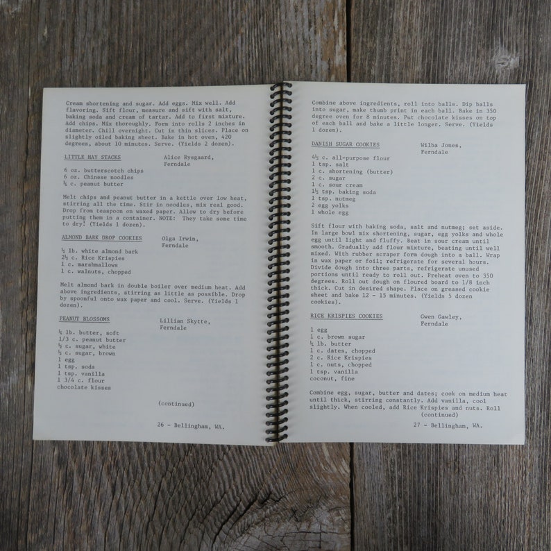 May include: A vintage cookbook with a brown leather cover and gold spiral binding. The pages contain handwritten recipes for various cookies, including peanut blossoms, almond bark drop cookies, little hay stacks, rice krispies cookies, and Danish sugar cookies.