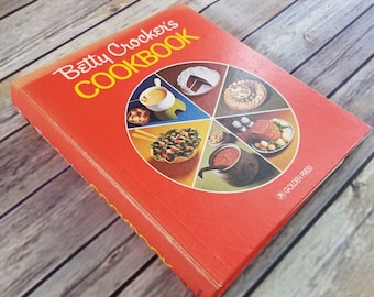 May include: A red hardcover cookbook titled "Betty Crocker's Cookbook" with a colorful illustration of various foods on the front cover. The illustration features a circle divided into six sections, each showcasing a different dish. The book is published by Golden Press.