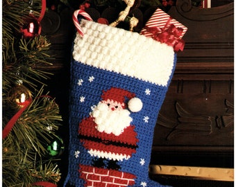May include: A blue and white crocheted Christmas stocking with a Santa Claus design. Santa is depicted climbing down a chimney with a red brick pattern. The stocking is hanging on a fireplace mantel with red candles and a Christmas card.
