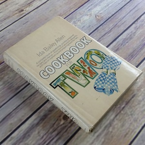 Puede incluir: Un libro de cocina blanco con el título "Cookbook for Two" en negrita, en negro. La portada presenta una ilustración colorida de la palabra "TWO" con una servilleta a cuadros.
