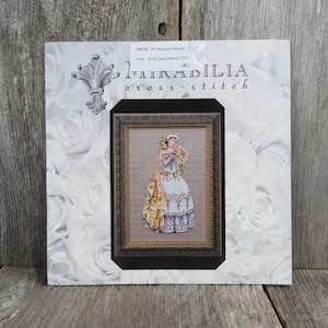 May include: Cross-stitch pattern featuring a woman in a yellow and white dress with a floral crown, holding a bouquet of flowers. The pattern is titled "The Blossom Harvest" and is from the brand "Mirabilia Cross-stitch".