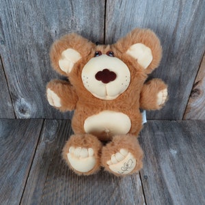 May include: A brown plush teddy bear with a light brown belly and a brown nose. The bear has a signature on one foot that says "Love, Bear".