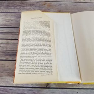 Pode incluir: Um livro aberto com uma capa amarela e p&aacute;ginas brancas. O texto na p&aacute;gina diz "Great Cookie News!" e discute receitas de biscoitos de um livro de culin&aacute;ria. O livro est&aacute; aberto em uma p&aacute;gina com um fundo branco e texto preto.