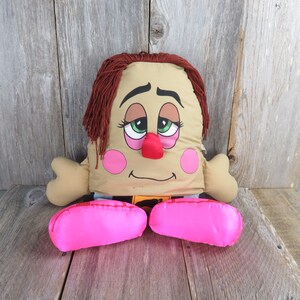 May include: A plush toy shaped like a person with a brown face, green eyes, a red nose, and pink cheeks. The toy is wearing blue pants and has pink feet. The toy has brown hair and a sleepy expression.