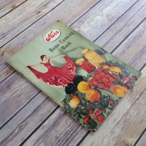 Puede incluir: Un libro vintage de conservas caseras Kerr de 1953 con una ilustración roja y blanca de una mujer con un vestido y frascos de fruta en conserva. La portada del libro presenta un fondo verde con una vid frondosa y bayas rojas.