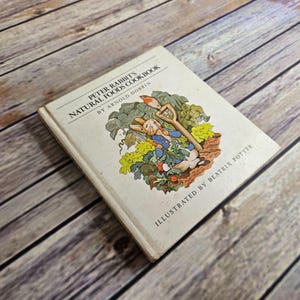 May include: A vintage cookbook titled "Peter Rabbit's Natural Foods Cookbook" by Arnold Dobrin, illustrated by Beatrix Potter. The cover features a colorful illustration of Peter Rabbit with a bird and a shovel, surrounded by foliage. The book is on a wooden surface.