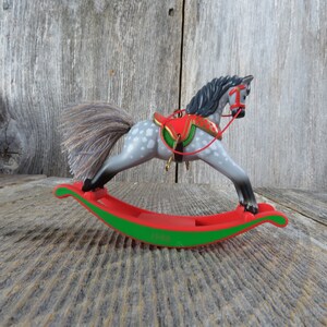 May include: A gray and white rocking horse Christmas ornament with a red saddle and green trim. The ornament is dated 1988.
