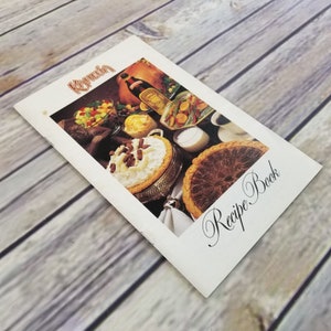 以下が含まれることがあります： 「Kona Koa Recipe Book」と黒色の筆記体で書かれた白いレシピ本。表紙には、ペカンパイ、白いトッピングのデザート、リキュールのボトルの写真が載っている。