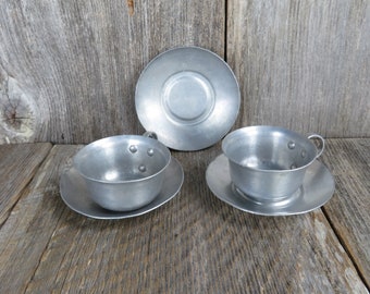 May include: A set of three silver-colored metal teacups and saucers. The cups and saucers are all the same size and have a simple, rounded design.