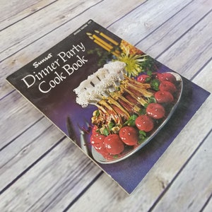 May include: A vintage Sunset Dinner Party Cookbook with a revised edition date of 1973. The cover features a colorful photo of a roasted meat dish with red apples and a white garnish.