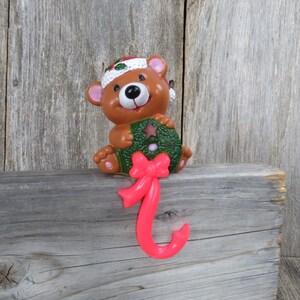Puede incluir: Un oso marrón con un gorro de Papá Noel sostiene una corona verde con un lazo rojo. El oso está unido a un gancho de media rosa.