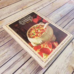 May include: A vintage cookbook titled "Woman's Day Encyclopedia of Cookery, Vol. 5." The cover features a bowl of food with a spoon, and the book is on a wooden surface. The book's title is in a decorative font.