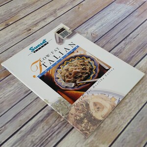 May include: A white cookbook with the title "Low-Fat Italian Cookbook" and a picture of a plate of pasta with a blue and yellow design. The book is part of the Sunset Books series and is for healthy eating.