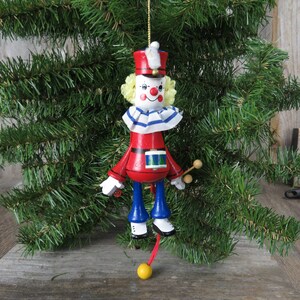 May include: A wooden Christmas ornament shaped like a clown with a red and blue uniform, a yellow hat, and a drum. The clown is holding drumsticks and has a yellow ball hanging from its feet.