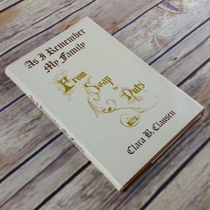Puede incluir: Un libro de tapa dura titulado "As I Remember My Family" y "From Soap to Nuts" de Clara B. Clausen. El libro tiene una cubierta blanca con letras doradas y granates y adornos decorativos. El libro está sobre una superficie de madera.