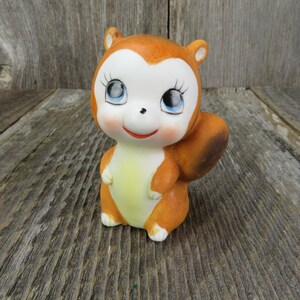 May include: A ceramic squirrel figurine with large, blue eyes and a brown and white body. The squirrel is standing on a wooden surface.