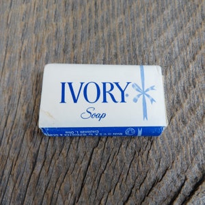 Puede incluir: Una barra de jabón Ivory blanca con letras azules y un diseño de flor azul. La barra está envuelta en papel blanco con texto azul que dice "Made in U.S.A. by Procter & Gamble, Cincinnati, Ohio."