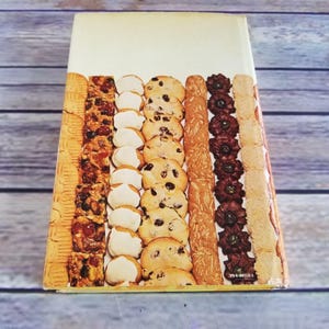 Pode incluir: Uma capa de livro de cozinha vintage que apresenta uma variedade de biscoitos e sobremesas, incluindo biscoitos de am&ecirc;ndoa, de gotas de chocolate e biscoitos amanteigados. A capa &eacute; amarela com uma borda marrom e o texto "39-4-8021" no canto inferior direito.