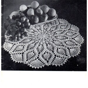 May include: A white crocheted doily with a scalloped edge, featuring a delicate floral design. The doily is displayed on a black surface with a silver bowl of fruit.