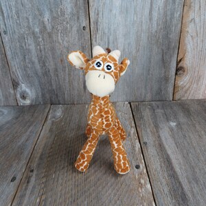 May include: A plush giraffe toy with a brown and white spotted pattern. The giraffe has large, round eyes, a cream-colored muzzle, and small brown horns. The toy is standing on a wooden surface.