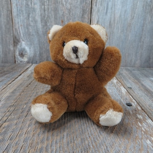 May include: A small brown teddy bear with a cream-colored nose and paw pads. The bear is sitting on a wooden surface.
