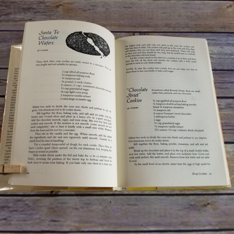 Pode incluir: Um livro de cozinha vintage aberto em duas p&aacute;ginas com receitas para "Santa Fe Chocolate Wafers" e "Chocolate Street Cookies". As receitas incluem ingredientes como manteiga, a&ccedil;&uacute;car, farinha e chocolate.