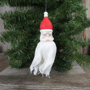 Puede incluir: Un adorno de Santa Claus de hilo blanco con un gorro rojo y ojos azules. El adorno está colgado de una rama de árbol.