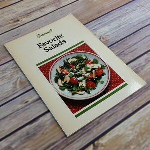 May include: A vintage Sunset magazine cookbook titled "Favorite Salads" with a photo of a salad on the cover. The salad is in a white bowl with a red and green border and contains mushrooms, tomatoes, olives, and greens.