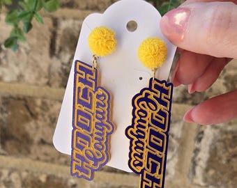 May include: Yellow pom-pom earrings with the text "HOOCH Cougars" in purple and gold. The earrings are hanging from a white card. The background is a brick wall.