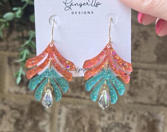 May include: Earrings in a leaf design, featuring orange and teal glitter resin, with a clear crystal charm. The earrings hang from gold-coloured hooks. The earrings are displayed on a white card with the text "Ginger Up DESIGNS".